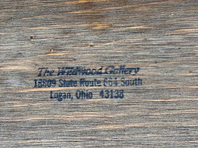 May include: Close-up of a wooden surface with the text "The Wildwood Gallery" and address details: 18809 State Route 664 South, Logan, Ohio 43138. The wood grain is visible.