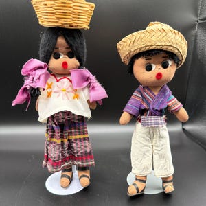 May include: Two handmade dolls. One doll has a woven basket on its head and wears a white blouse with pink accents and a patterned skirt. The other doll wears a straw hat, a purple and white striped shirt, and white trousers. Both dolls have black hair and sandals.
