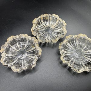 May include: Three clear glass dishes with a scalloped, flower-like edge and a radial ribbed pattern radiating from the center. Each dish features a transparent, reflective surface with subtle gold-toned highlights along the outer scalloped rim.