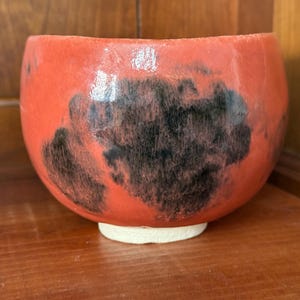 May include: A handmade ceramic bowl with a vibrant red glaze and dark, mottled black accents. The bowl has a rounded shape and sits on a small, off-white base. This unique piece is perfect for serving or display.