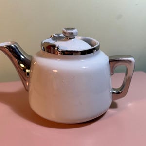May include: A white teapot with a silver lid and handle. The teapot is small and has a rounded body.