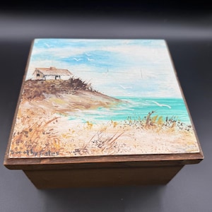 May include: A wooden box with a painted coastal scene on the lid. The artwork depicts a small white building on a hill overlooking the ocean, with blue sky and seagulls. The artist's signature, "Dorothy Gates", is visible.