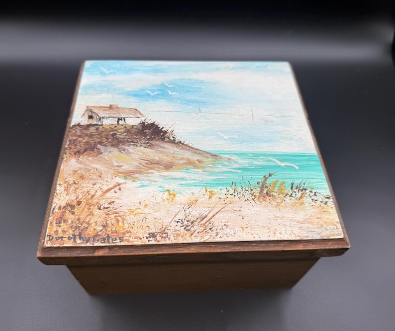 May include: A wooden box with a painted coastal scene on the lid. The artwork depicts a small white building on a hill overlooking the ocean, with blue sky and seagulls. The artist's signature, "Dorothy Gates", is visible.