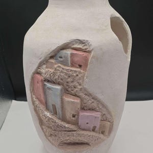 May include: Off-white ceramic vase with a carved relief of a village scene. The relief features small, colorful buildings in shades of pink, blue, and tan. The vase has an oval shape and a large opening on the side.
