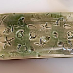 May include: A rectangular, ceramic serving tray with a sea life theme. The tray is glazed in shades of green and features embossed starfish, seashells, and spiral shell designs. The edges are rounded, and the overall design has a rustic, handmade appearance.