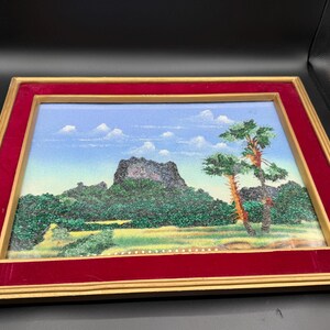 May include: A rectangular framed landscape artwork featuring a textured mountain scene with green foliage and two prominent trees under a blue sky with white clouds. The frame consists of a gold-toned outer border and a deep red velvet inner mat.