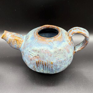 May include: A handmade ceramic teapot with a round body, a spout, and a handle. The teapot has a blue and brown glaze with a textured surface. The interior is dark blue.