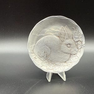 May include: A silver-colored, circular decorative plate with a carved design of a rabbit surrounded by leaves. The plate is supported by a clear, angled stand, and is set against a dark background.