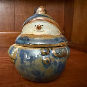 May include: A ceramic snowman-shaped container with a lid. The snowman has a blue hat and body with brown and tan accents. The face has a small brown nose and black eyes. The container is sitting on a wooden surface.