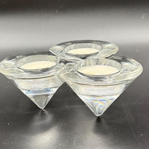 May include: A clear glass candle holder with three diamond-shaped sections. Each section holds a small, white candle. The candle holder is designed to hold tea lights and is suitable for home decor.