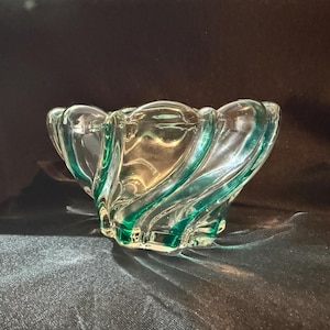 Peut inclure: Un bol en verre transparent avec un motif tourbillonnant et des accents turquoise. Le bol a une forme unique avec un bord festonné. Le verre est transparent, laissant passer la lumière, et est posé sur un fond sombre.