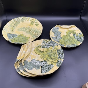 May include: Three ceramic plates with irregular, wavy edges. The plates feature a pale yellow background with dark green botanical and geometric patterns, including fern leaves, succulent-like flowers, and honeycomb shapes printed on the surface.