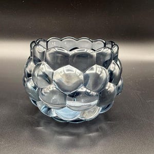 May include: A blue glass votive candle holder with a textured, bubble-like design. The candle holder has a round shape and a scalloped rim. The glass is transparent, allowing light to pass through.