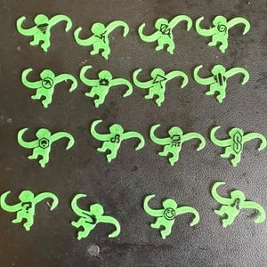 May include: A collection of fourteen small, light green, monkey-shaped cutouts arranged in four rows. Each monkey figure features a unique black graphic symbol printed on its chest, including geometric shapes, letters, and a smiley face icon.
