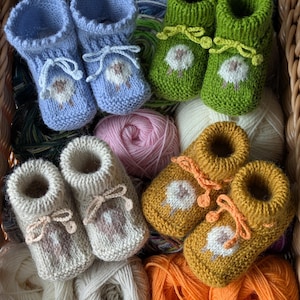 Pode incluir: Uma cesta cheia de botinhas de bebê feitas à mão em tricô em várias cores: azul claro, verde, bege e amarelo mostarda. Cada par apresenta um desenho de ovelha e laços de fita. A cesta também contém novelos de lã em cores coordenadas.