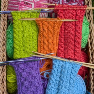 May include: A wicker basket filled with multiple balls of yarn in pink, green, orange, and blue, featuring four rectangular knitted swatches in lime green, orange, red, and purple, each attached to wooden or metal knitting needles.