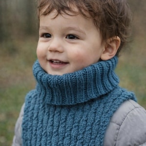 Könnte beinhalten: Ein tealblauer Strickschal mit einem strukturierten Zopfmuster. Der Schal wird von einem Kind getragen, kombiniert mit einer grauen Jacke. Der Hintergrund ist verschwommen und deutet auf eine Umgebung im Freien hin.