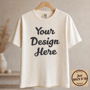 May include: Cream-colored t-shirt with the text "Your Design Here" in a bold, black font. The shirt is hanging on a wooden hanger. A small logo is visible on the upper right corner of the shirt. The shirt is a Comfort Colors brand.