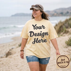 May include: A person wearing a light yellow t-shirt with the text "Your Design Here" in black. The person is wearing a brown cap and denim shorts. A small sign with the text "Just MOCK It Up SHOP" is held in the hand.