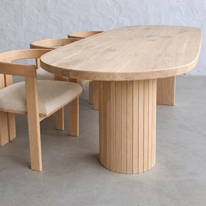 Può includere: Un tavolo da pranzo ovale in legno chiaro con una spessa base cilindrica a doghe verticali. Il tavolo è accompagnato da diverse sedie in legno chiaro abbinate, dotate di schienali curvi e cuscini di seduta in tessuto testurizzato color bianco sporco.