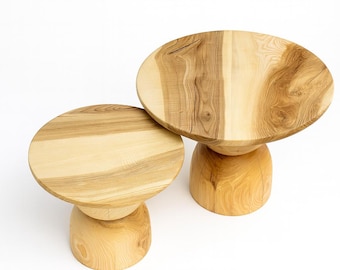 Puede incluir: Dos mesas auxiliares de madera con acabado natural. Cada mesa tiene una parte superior redonda y una base de pedestal redondeada. La veta de la madera es visible, mostrando la belleza natural del material. Las mesas son de color marrón claro.
