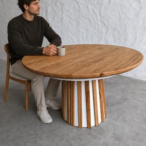 以下が含まれることがあります： 白と木製のスラットベースを備えた丸い木製ダイニングテーブル。テーブルトップは明るい茶色で、木目がはっきりと見えます。テーブルの直径は約122cmです。人がテーブルに座っています。