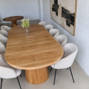 Op de afbeelding: Een ovale houten eettafel met een centrale kolomvoet, omringd door tien gebroken witte gestoffeerde stoelen met zwarte metalen poten. Op het tafelblad met zichtbare houtnerf staat een doorzichtige glazen vaas met groene bloemen.