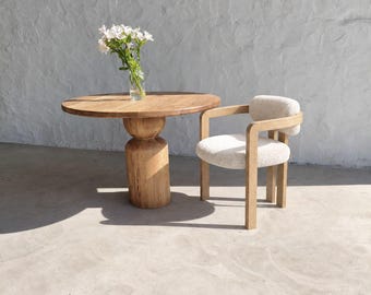 Op de afbeelding: Een ronde houten tafel met een dikke kolomvoet en een bijpassende houten stoel met een crèmekleurige, gestructureerde bekleding op de zitting en rugleuning. Een doorzichtige glazen vaas met witte bloemen staat op de tafel.