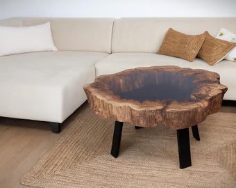 May include: A unique, live-edge coffee table with a dark, glossy center. The table is made of natural wood with a dark brown finish and sits on three black metal legs. The table is placed on a woven jute rug in front of a cream-colored sectional sofa.