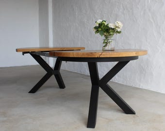 May include: A dining table with a round, light brown wooden top and a black metal base featuring X-shaped legs. A clear glass vase containing white hydrangea flowers sits on the surface of the table, which is partially extended.
