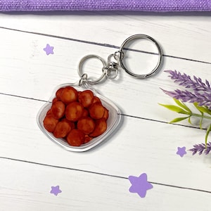 May include: A clear acrylic keychain featuring a bowl of reddish-brown food items. The keychain has a silver metal ring and clasp. The background includes purple star-shaped confetti and a sprig of lavender on a white wooden surface.