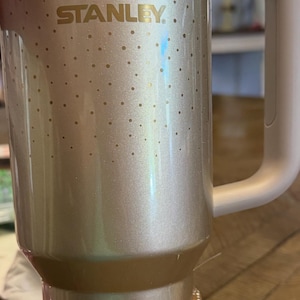 May include: A gold-coloured Stanley flask with a handle. The flask has a gold logo and the word "STANLEY" printed on the side. The bottom is decorated with gold and silver rhinestones and beads.