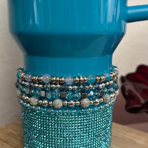 May include: A turquoise mug with a beaded wrap. The wrap features layers of silver beads, iridescent beads, and various shades of blue beads. The bottom of the mug is covered in a sparkling turquoise material.