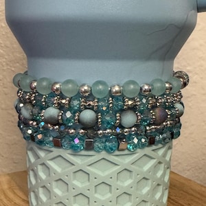 May include: A stack of beaded bracelets in shades of blue and silver. The bracelets are displayed on a light blue tumbler with a geometric pattern. The bracelets feature various bead sizes and textures, creating a layered look.