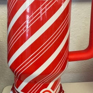 May include: Red and white striped Stanley tumbler with a red handle. The tumbler has the brand name "STANLEY" printed on it. The bottom of the tumbler is decorated with beaded jewelry.