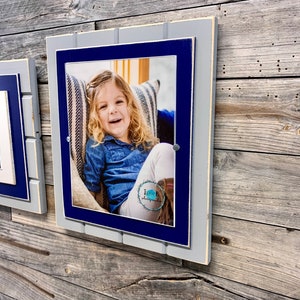 May include: A gray and blue wooden picture frame with a photo of a young girl smiling. The frame has a white border and is hanging on a wood wall.