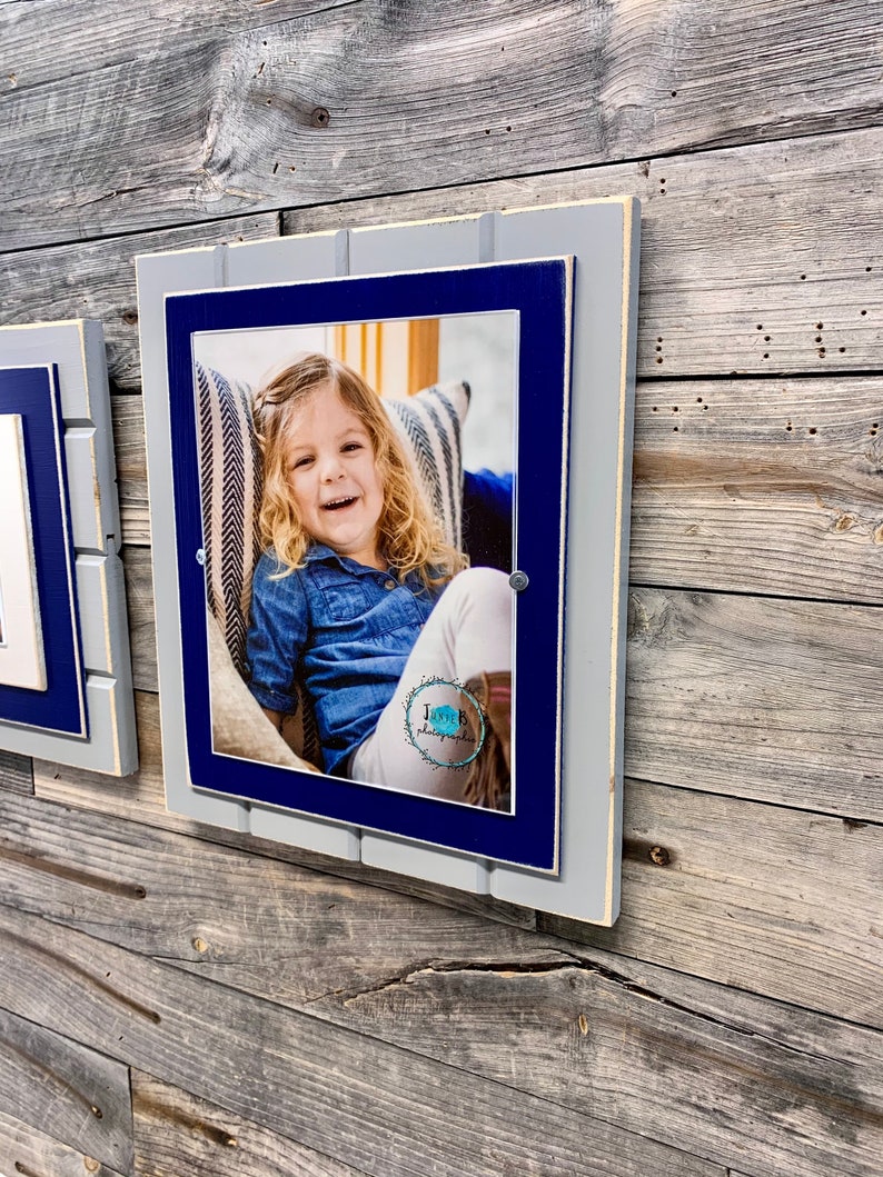 May include: A gray and blue wooden picture frame with a photo of a young girl smiling. The frame has a white border and is hanging on a wood wall.