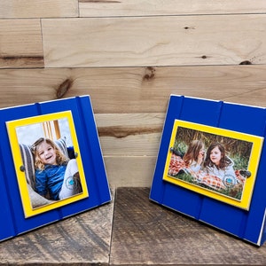May include: Two blue and yellow wooden picture frames with a white border. The frames are standing upright on a wooden surface. The frames have a photo of a young girl in one and two young girls in the other.