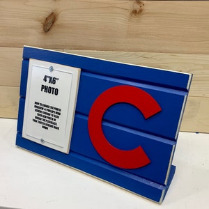 May include: A blue and white wooden sign with a red "C" in the center. The sign has a white frame with the text "4"x"6" Photo" and instructions on how to change the photo.