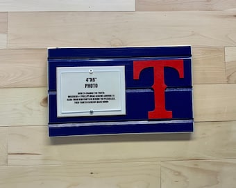 May include: A blue and white wooden sign with a red "T" in the center. The sign has a white frame with the text "4"x6" Photo" and instructions on how to change the photo.