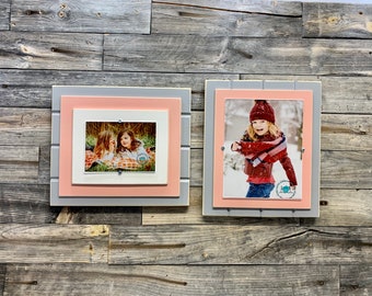 May include: Two wooden picture frames with a light gray outer frame and a pink inner frame. The frames are hanging on a gray wood wall. One frame has a photo of two girls in a field, the other frame has a photo of a girl in a winter scene.