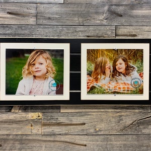 May include: Two black picture frames with white mounts, each displaying a photograph of children. The left frame shows a young girl with curly hair, while the right frame shows two girls lying on a blanket.