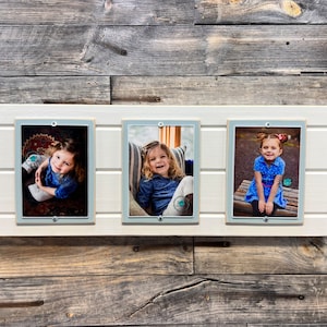 May include: Three white picture frames with a light blue border, each containing a photo of a young girl. The frames are mounted on a white wooden board.