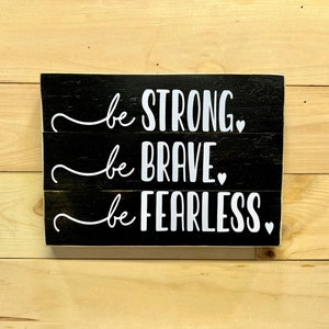 Puede incluir: Un letrero de madera negro con letras blancas que dice "be STRONG, be BRAVE, be FEARLESS". El letrero está hecho de tres tablones horizontales y tiene un estilo rústico y de granja. El fondo es de madera clara.