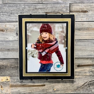 May include: A framed photograph featuring a young person in winter attire, smiling in the snow. The frame has a black outer border, a gold inner border, and is mounted on a rustic wooden background. The photograph is signed by "Junte B Photography".