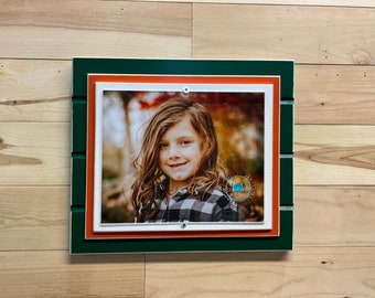 May include: A green, orange, and white picture frame with a photo of a young girl with long brown hair smiling. The photo is set against a blurred background of fall foliage.