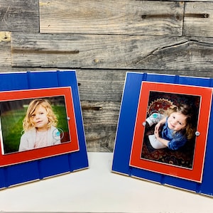 May include: Two blue and red wooden picture frames with white accents. Each frame holds a photo of a young girl. The frames are standing upright on a wooden surface.