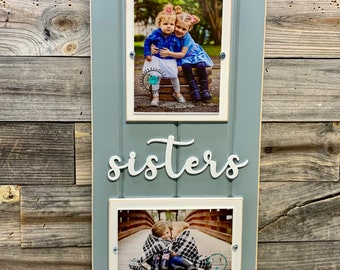 May include: A light blue wooden picture frame with two white picture frames inside. The top frame has a photo of two young girls hugging. The bottom frame has a photo of two young girls hugging on a bridge. The word "sisters" is written in white letters on the frame.