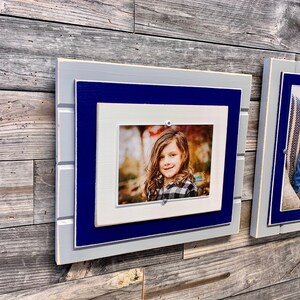 May include: A set of two picture frames with a rustic wood look. The frames are layered with a white, navy blue, and gray paint. The frames are hanging on a wood wall.