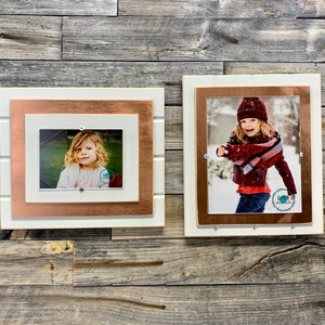 May include: Two framed photos of children, one in a white frame with a copper inner frame and the other in a white frame with a brown inner frame. Both frames have a rustic woodgrain finish.